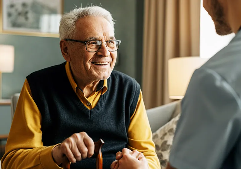 Ein älterer Mann mit grauem Haar und Brille sitzt lächelnd in einem gemütlich eingerichteten Raum und hält einen Gehstock in der Hand. Er trägt ein senfgelbes Hemd und eine dunkle Strickweste, was ihn warm und freundlich wirken lässt. Vor ihm sitzt eine Person, deren Hand er freundlich hält, was eine vertraute, herzliche Verbindung vermittelt. Im Hintergrund sind weiche Beleuchtung und Vorhänge zu sehen, die dem Raum eine ruhige und einladende Atmosphäre verleihen. Die Szene strahlt Zufriedenheit und Wohlbefinden aus und repräsentiert die persönliche und respektvolle Betreuung von SANICUR Dresden GmbH.