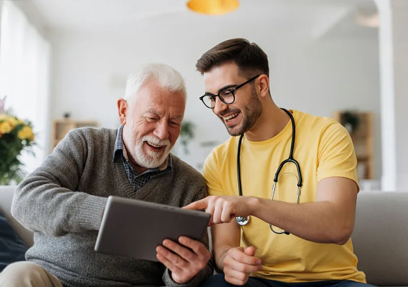 Ein junger Mitarbeiter der SANICUR Dresden GmbH, erkennbar an einem Stethoskop um den Hals, sitzt neben einem älteren Mann und zeigt ihm lächelnd etwas auf einem Tablet. Der ältere Mann mit weißem Haar und Bart lacht und schaut interessiert auf das Gerät. Die beiden sitzen in einem gemütlichen Raum mit Blumen im Hintergrund, und die entspannte, freundliche Atmosphäre vermittelt Freude und Offenheit im Umgang mit moderner Technik für die Unterstützung älterer Menschen. Das Bild symbolisiert digitale Unterstützung und gemeinsame Interaktion in der Pflege.